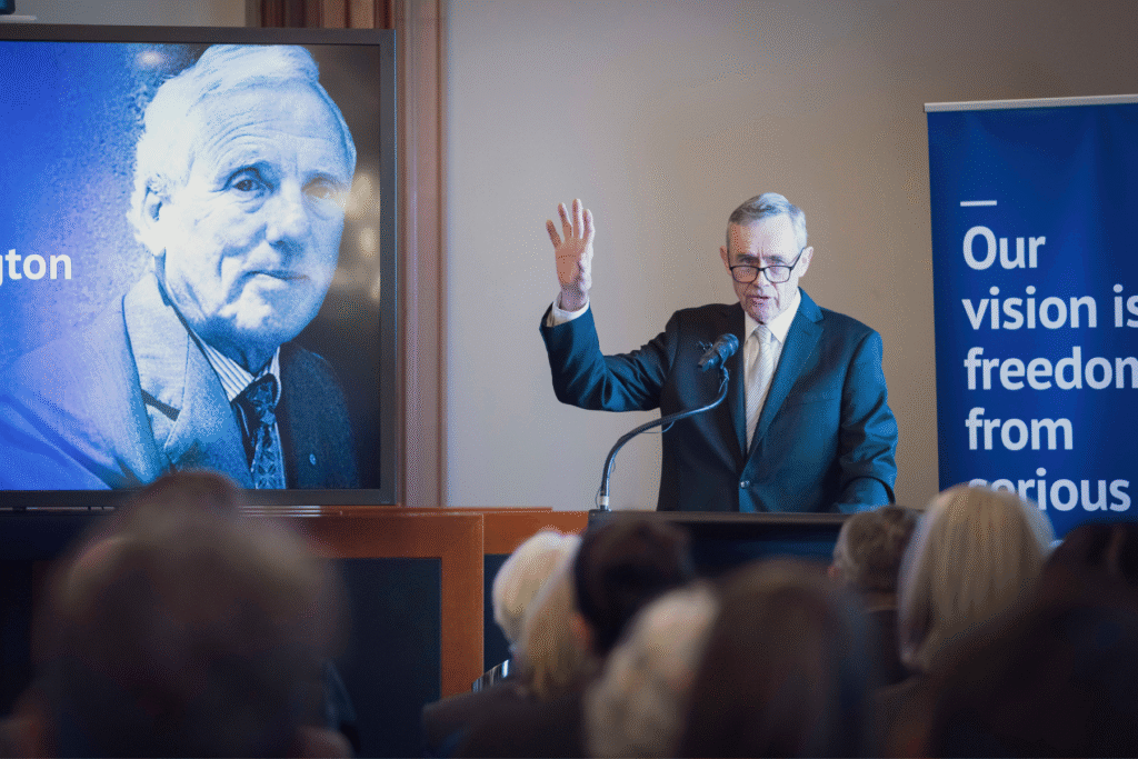 His Honour Anthony Parsons addresses audience from lectern and mic, behind him to the left is a screen with David Penington's face on it
