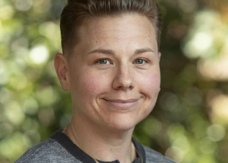 This shows a smiling person with short hair, a grey top and a gold chain.